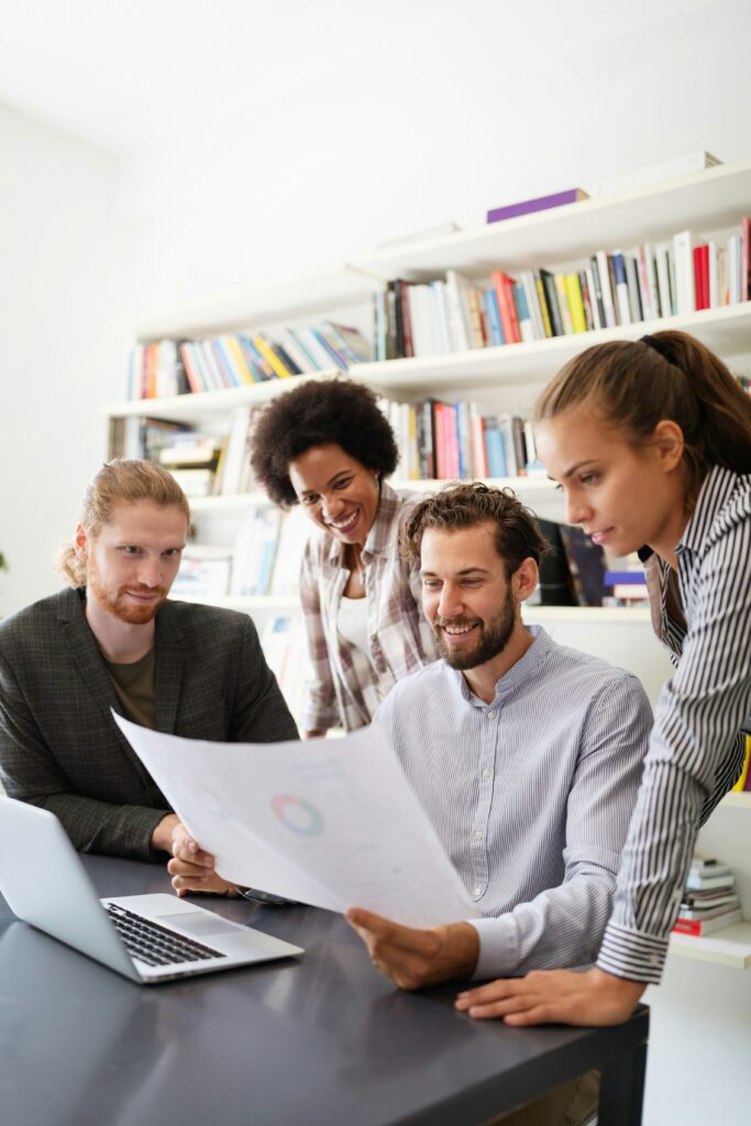 Four coworkers gather around a table in a bright office, reviewing a printed document together beside an open laptop.